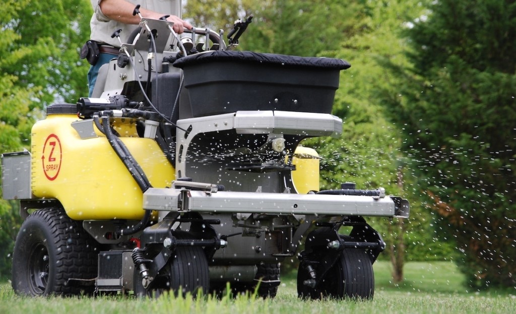 lawn care professional operating a stand-on spreader sprayer, applying granular fertilizer or herbicide to a green lawn